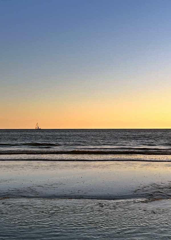 Bølgerne skvulper over strandkanten, sejlbåden tegnes flot mod den flotte nedgående sols himmel